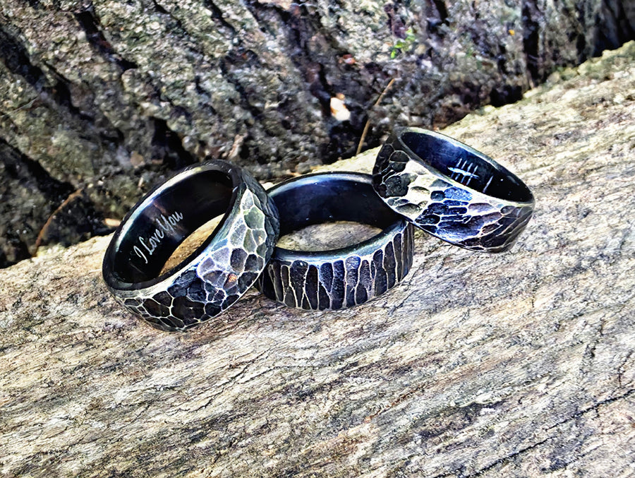 Three hammered metal rings with a dark oxidized finish and textured outer bands, resting on a piece of weathered wood outdoors.