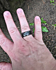 A close-up of a hand wearing a dark rectangular ring with a hammered metallic pattern, set against a background of dark soil and small green plants.