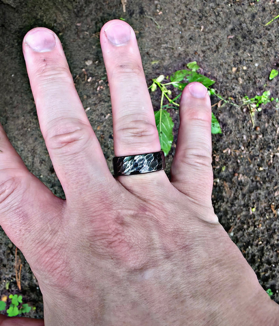 A close-up of a person's hand wearing a wide black band ring with a hammered or textured metallic surface, resting on dark soil with small green plants.