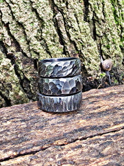 Three stacked hammered metal rings with a dark oxidized finish and textured organic patterns, displayed on a rustic log in front of a mossy tree trunk.