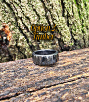 A textured dark metal ring with a bark-like pattern sits on a weathered log with tree bark in the background. Yellow text overlay reads 'Option 2: Timber'.