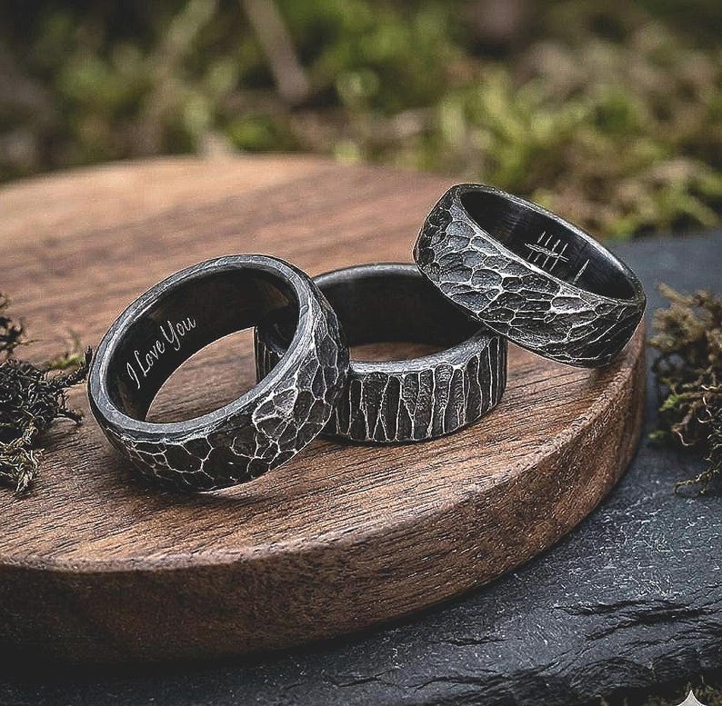 Three dark metallic rings with textured, hammered surfaces and engraved details, displayed on a round wooden board with mossy props in a blurred outdoor setting.