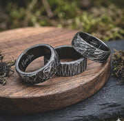 Three dark metallic rings with textured, hammered surfaces and engraved details, displayed on a round wooden board with mossy props in a blurred outdoor setting.