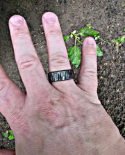 A close-up of a person's hand wearing a textured black band ring outdoors on dark soil with small green plants.